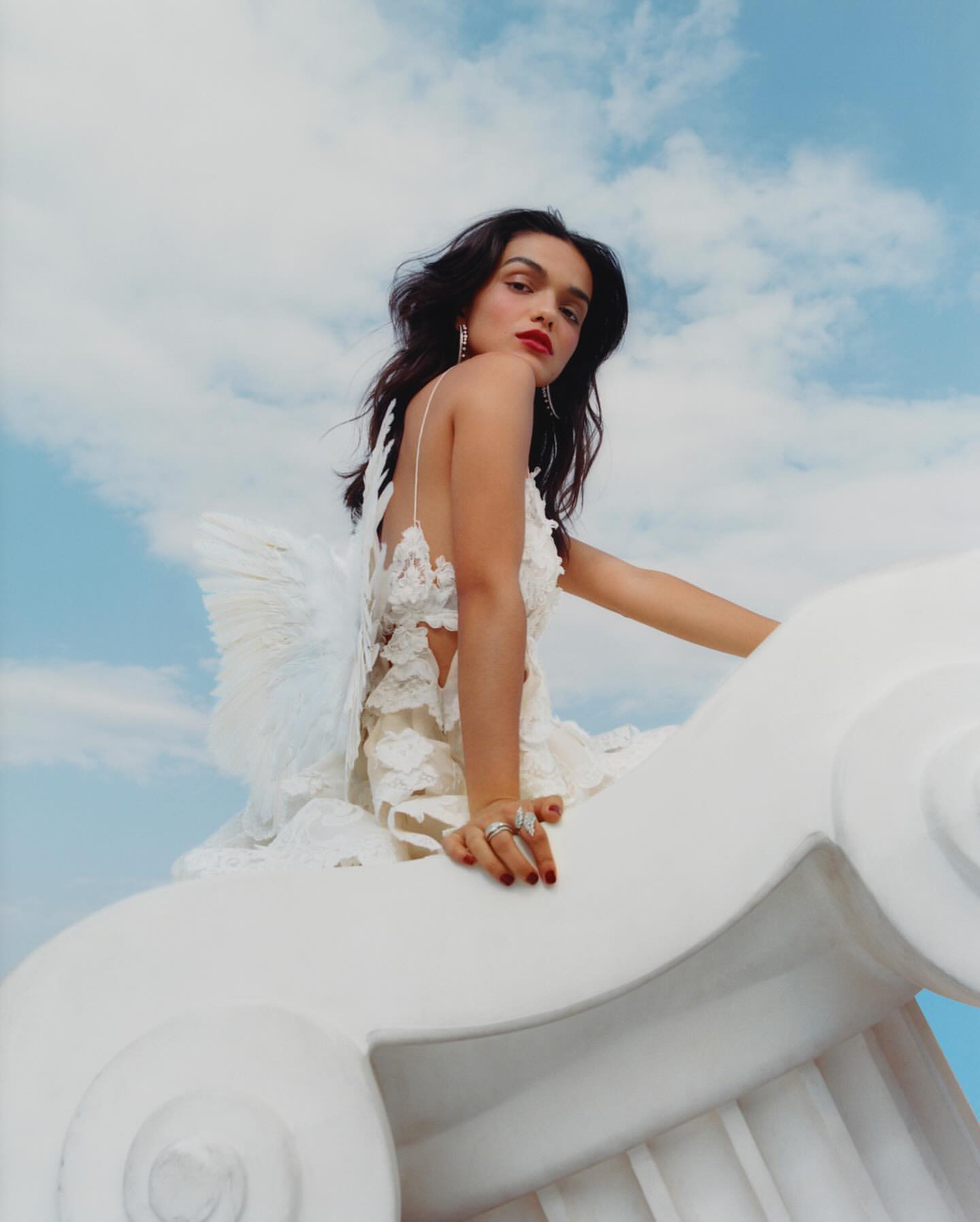 Young woman in angel costume with white dress and wings posing on a white sculpture against a blue cloudy sky background.