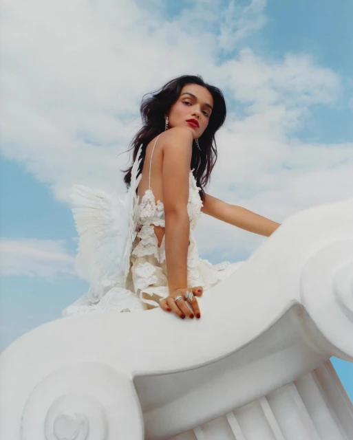 Young woman in angel costume with white dress and wings posing on a white sculpture against a blue cloudy sky background.