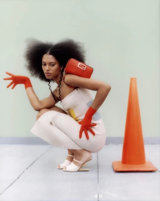 Stylish woman in orange gloves poses in a squat wearing a white dress with a matching orange purse and an orange traffic cone.