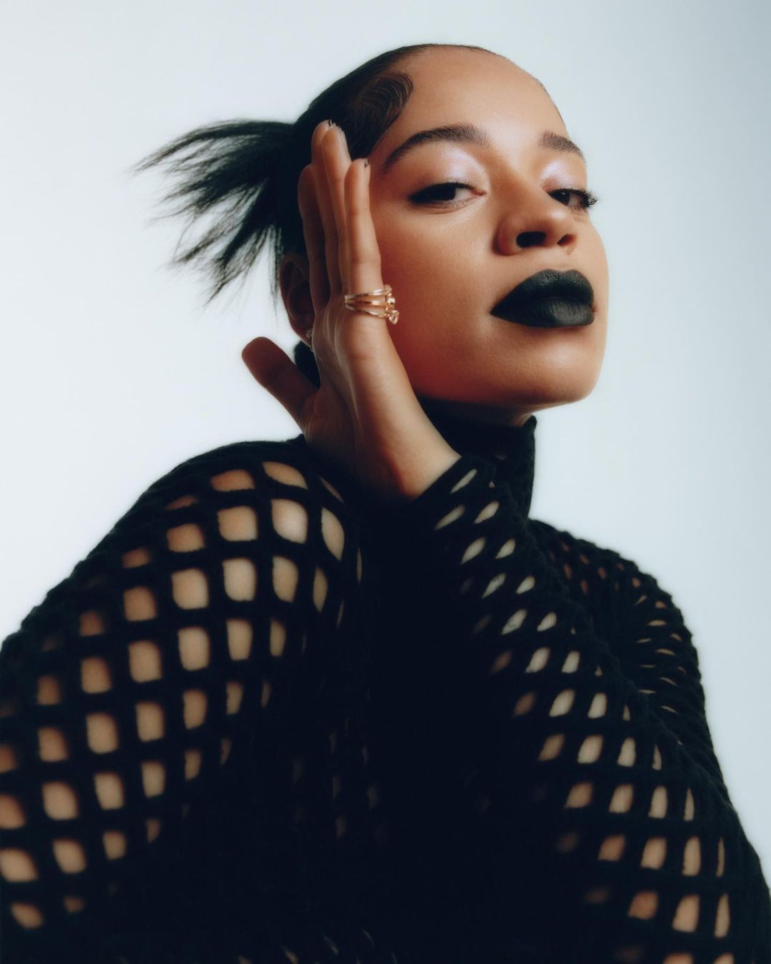 Portrait of a woman with dark lipstick wearing a black mesh top, posing in front of a bright white background in a studio setting.
