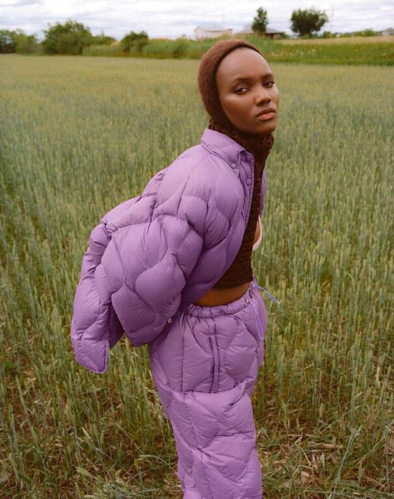 Adult woman wearing a purple quilted jacket and pants poses outdoors in a field of wheat. Fashion shot in a natural environment.