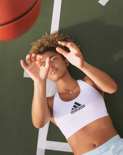 Athletic woman in white sports bra looks at basketball overhead on green court with white lines; Adidas logo visible on her sports bra.