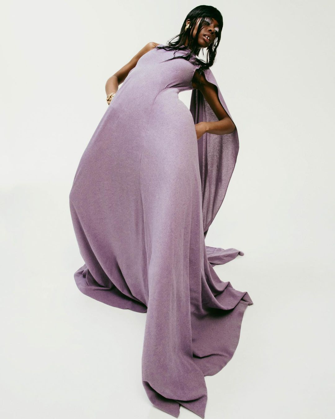A Black female model in a flowing lavender gown poses against a white backdrop in a studio setting, showcasing fashion and beauty.
