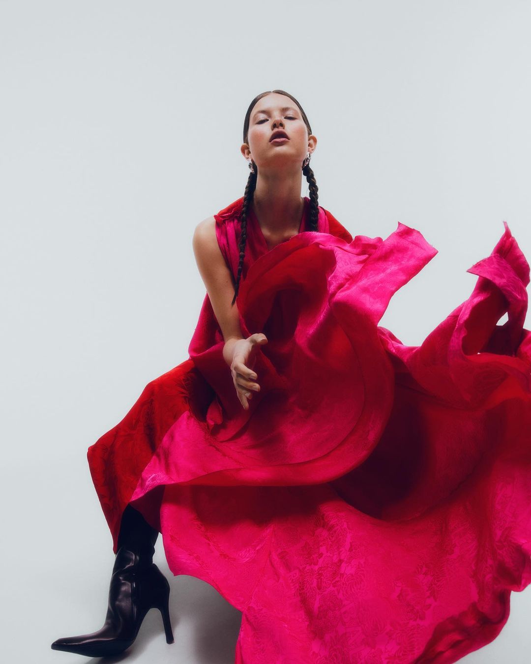 Elegant adult female in a pink gown and black high-heeled boots poses in a photo studio against a white backdrop.