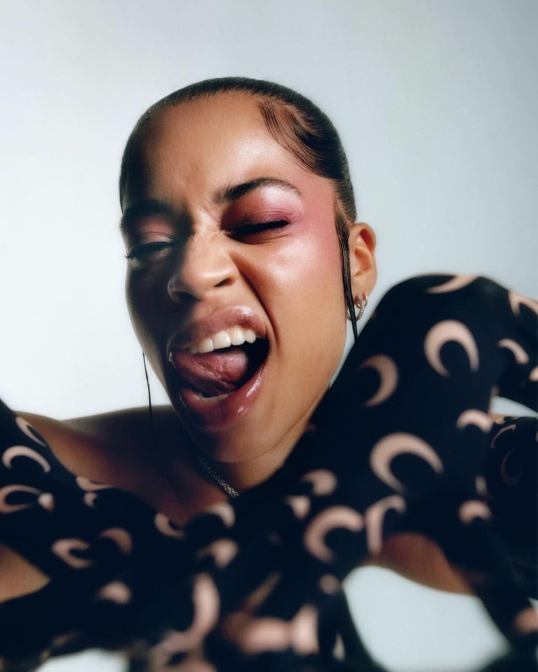 Close-up portrait of a woman with pink makeup and patterned sleeves, sticking her tongue out against a bright background.