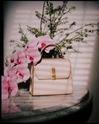A structured pink handbag resting on a marble tabletop next to cascading pink orchid flowers, photographed with soft window blind shadows.