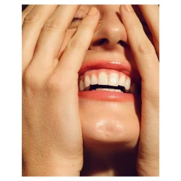 Close-up portrait of a person holding their hands up to frame their laughing face, showing teeth