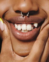Close-up portrait of a smiling person holding their cheeks, featuring a silver septum piercing and manicured nails.