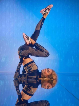 Model performing a flexible yoga pose wearing patterned athletic wear against a bright blue background with a glossy floor reflection.