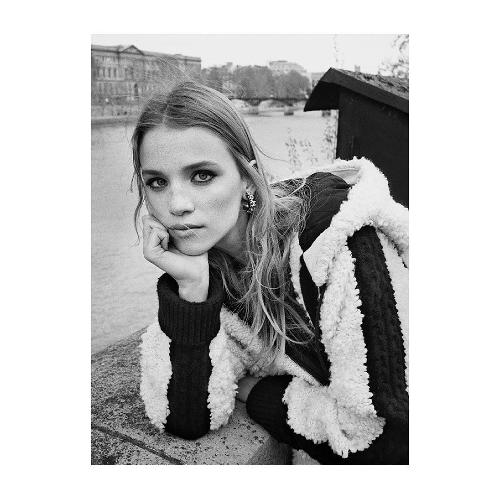 Black and white portrait of a young woman with long hair leaning her head on her hand outdoors with a bridge and building in the background.