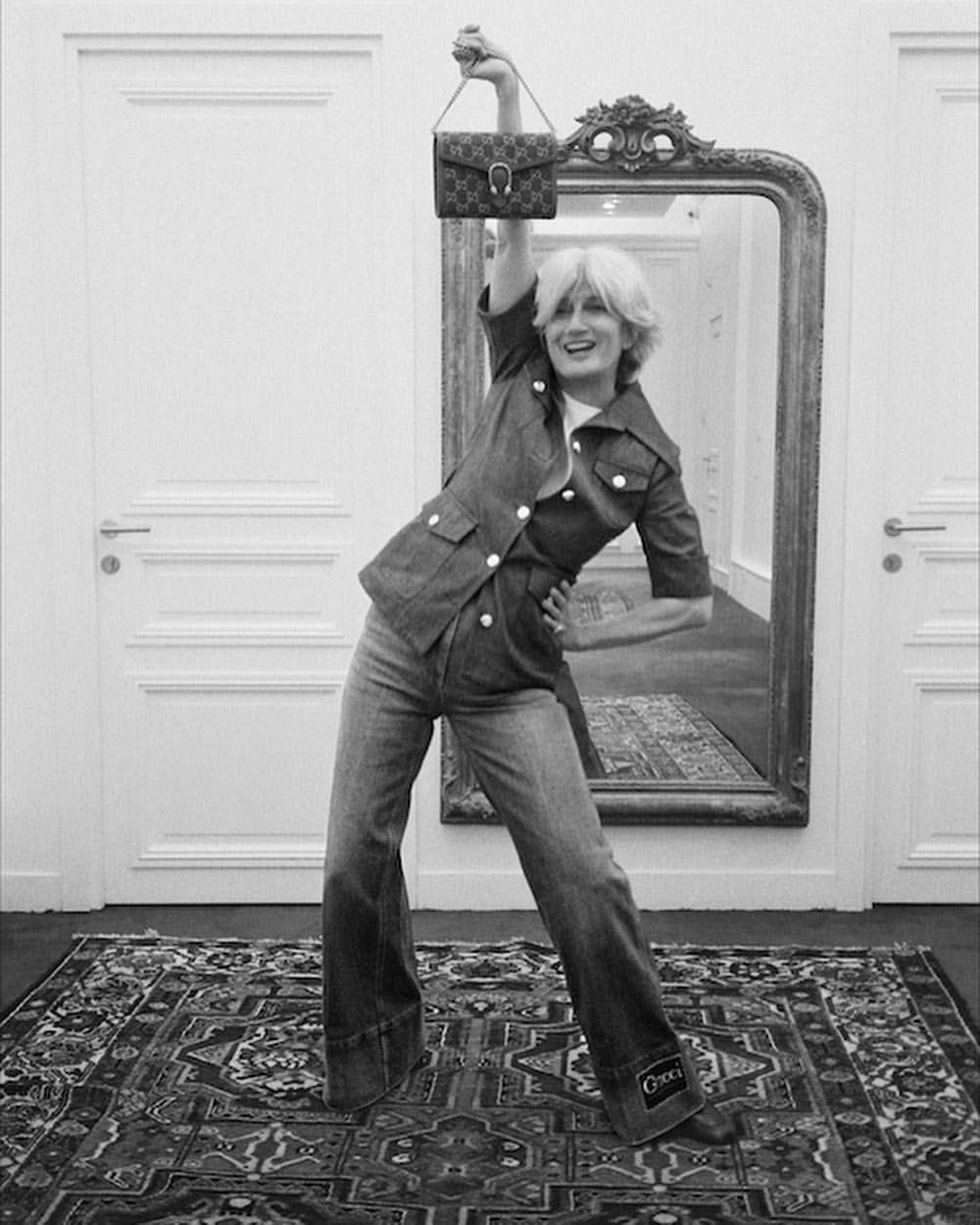 A woman poses with a purse indoors, wearing a denim jacket and bell-bottom jeans, on a patterned rug with doors and a mirror behind her.
