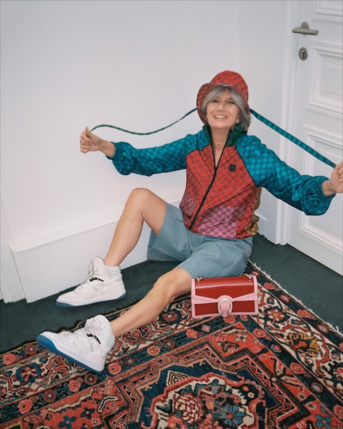 Older female fashion model sits on an ornate rug indoors, wearing a colorful jacket, shorts, sneakers, bucket hat, and handbag.