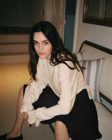 Brunette woman in beige sweater and black skirt sitting on a sofa in an indoor setting, posing for a portrait-style photograph.