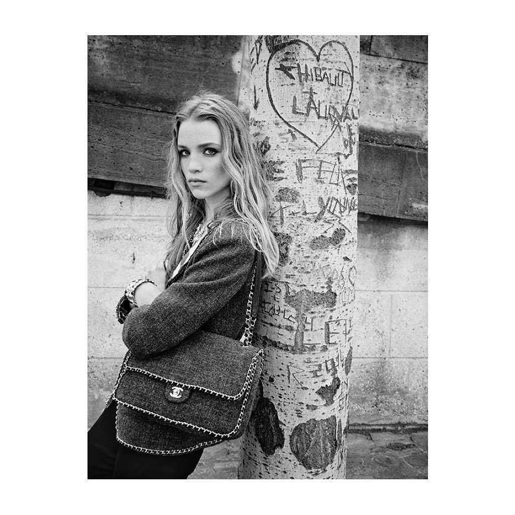 A blonde woman in a blazer stands near a tree trunk in an urban setting with a handbag, captured in a black and white medium shot.