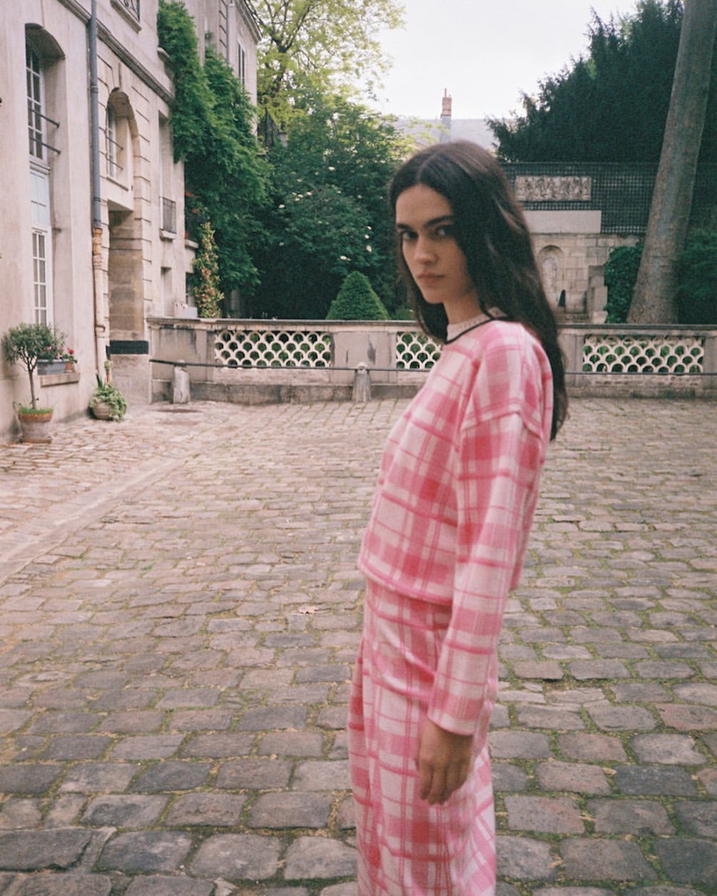 Brunette woman in pink plaid matching set poses outdoors in courtyard with stone pavers and historic building in the background on an overcast day.