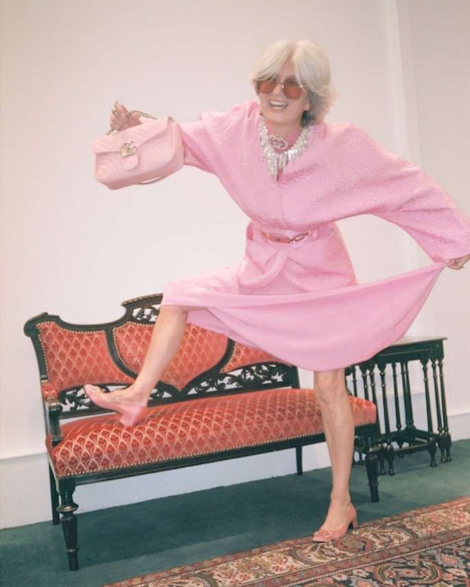 Elegant older woman in a pink dress and sunglasses stands on a red and gold settee holding a matching pink purse, smiling.
