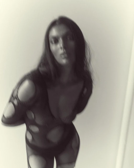 Monochrome image of a woman in a fishnet bodysuit posing in front of a plain wall with soft lighting, capturing a fashion forward look.