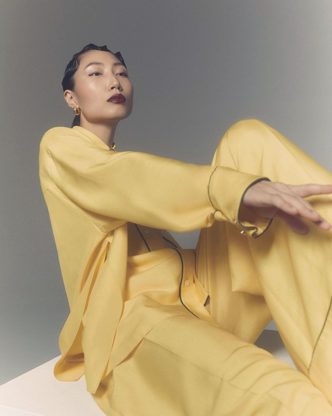 Elegant Asian woman in yellow pyjamas sits in studio, gazing up. Chic, short, slicked-back hair and dark lipstick enhance the image.