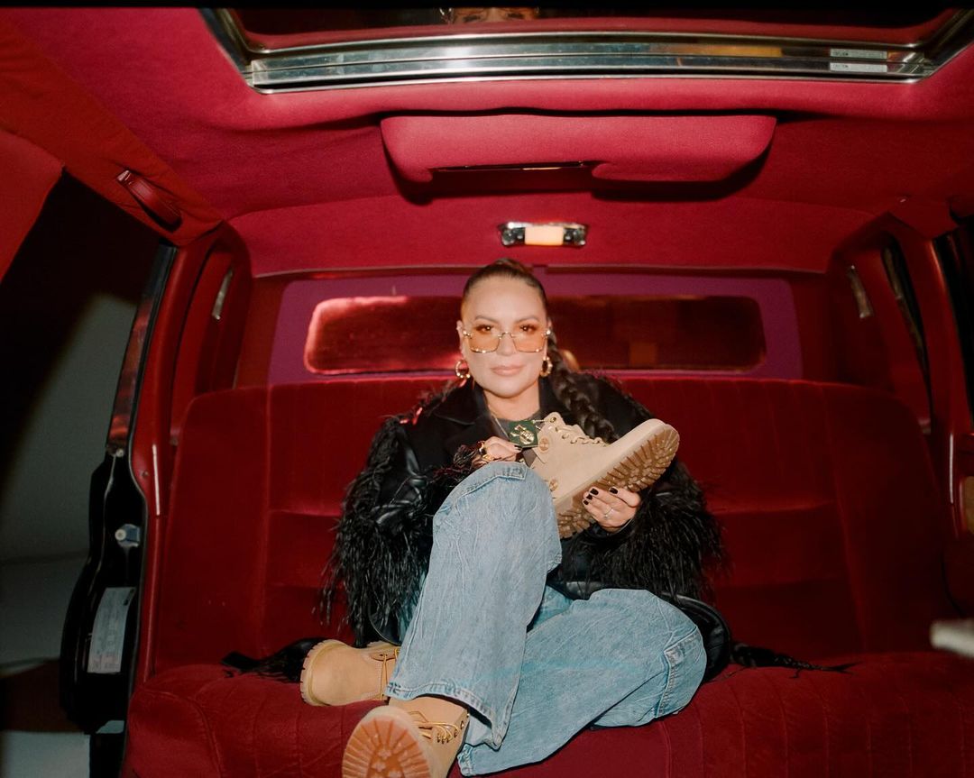 Woman in a car interior holds a tan Timberland boot, wearing jeans, jacket with feathers, and glasses, against a red velvet backdrop.