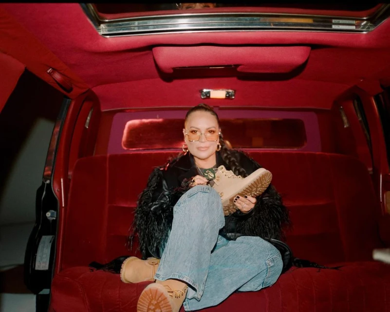 Woman in a car interior holds a tan Timberland boot, wearing jeans, jacket with feathers, and glasses, against a red velvet backdrop.