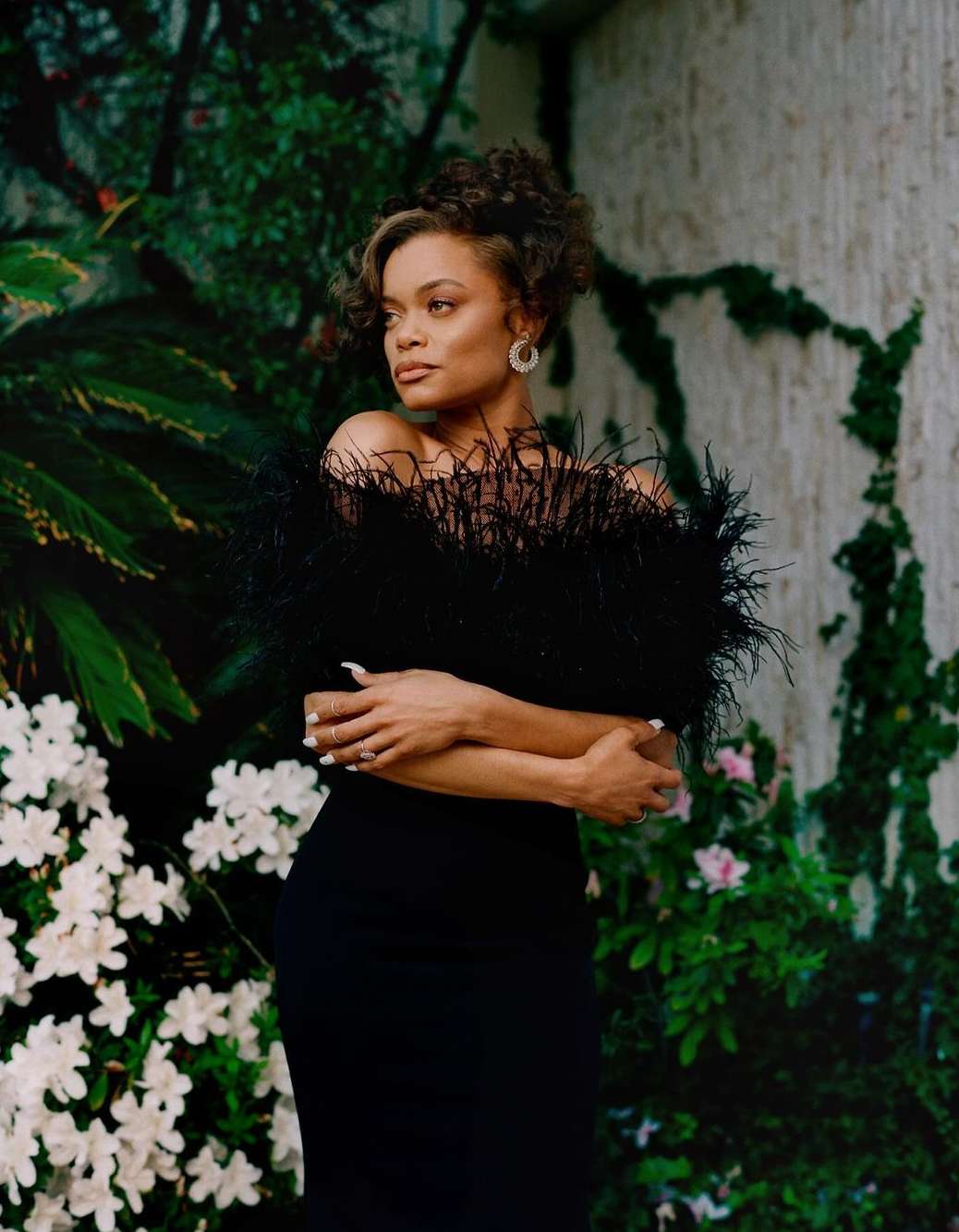 Elegant woman in a black dress poses against a lush green background with white flowers, creating a striking contrast of colors and textures.