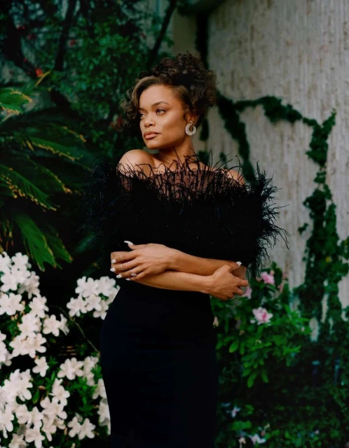 Elegant woman in a black dress poses against a lush green background with white flowers, creating a striking contrast of colors and textures.