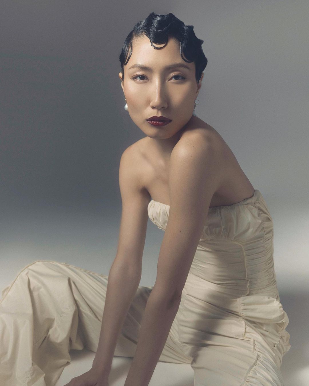 Portrait of an Asian woman with short, waved black hair, wearing a strapless off-white dress and pearl earrings, posing against a grey background.