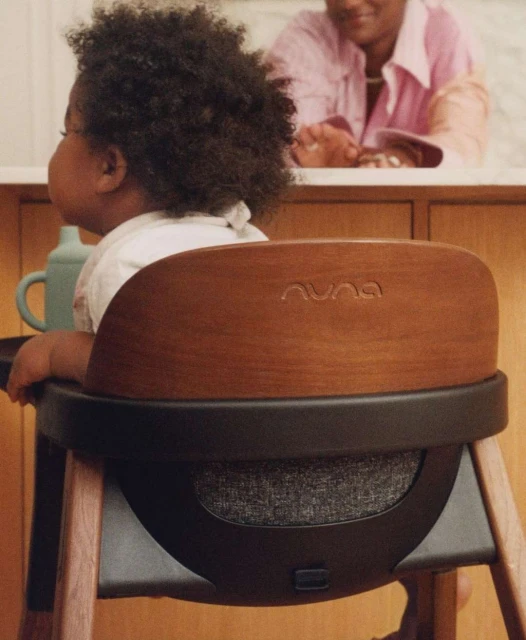 Child sits in Nuna high chair with wood accents and black frame, adult in background. Focus on baby and chair. Indoor setting.