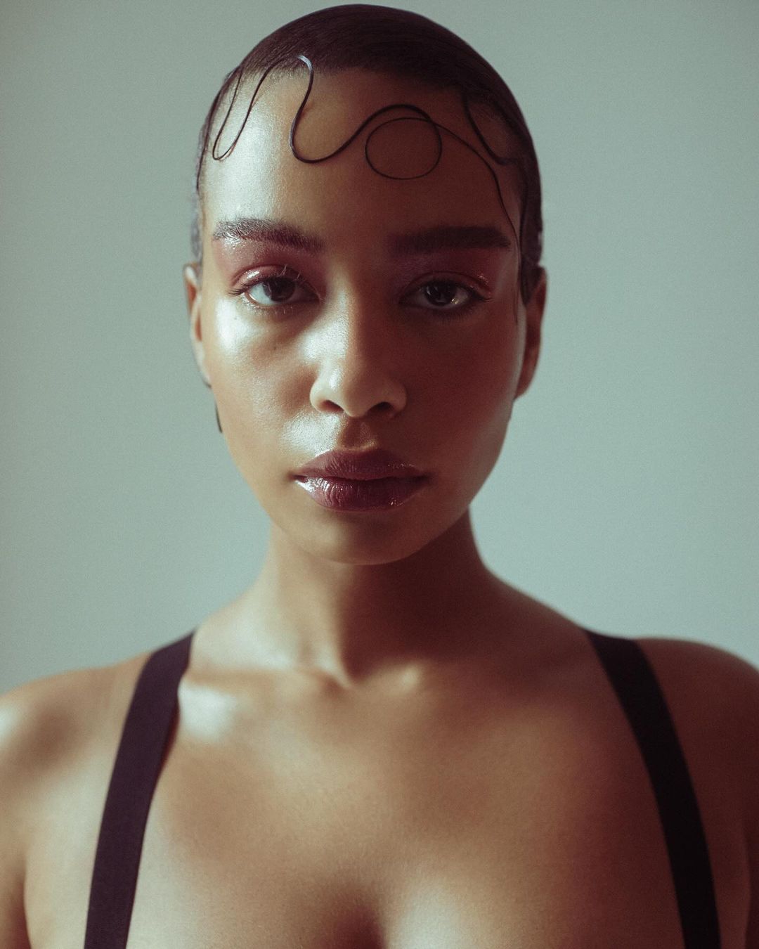 Close-up studio portrait of a woman with glossy makeup and slicked-back hair, wearing black straps, against a neutral background.