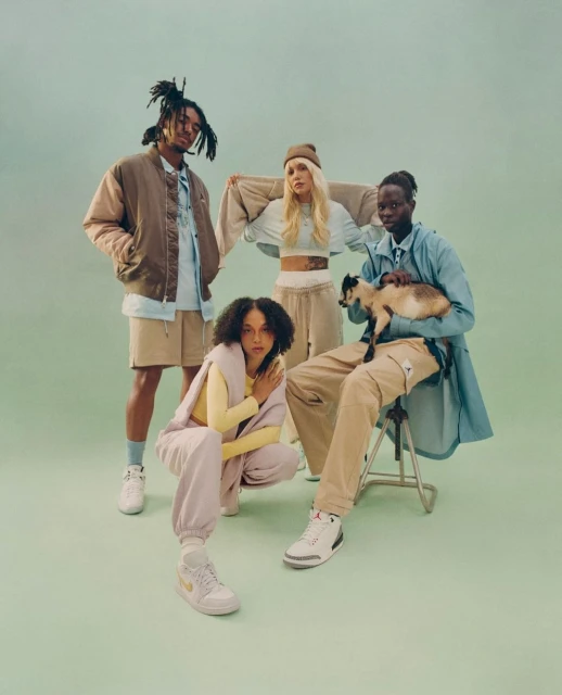 Group of four adult fashion models in casual clothing posing in a studio with soft, diffused lighting and a solid light green backdrop.