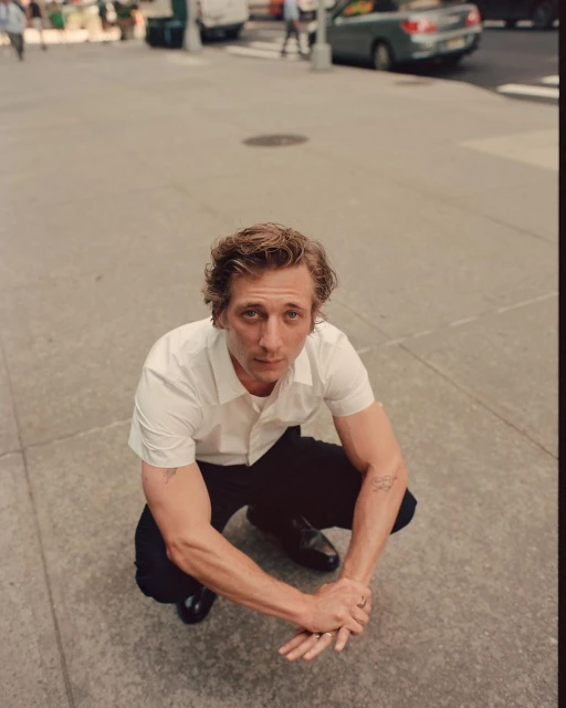 Adult male in a white short-sleeve shirt and black pants crouches on the sidewalk in an urban environment during daylight hours, looking up.