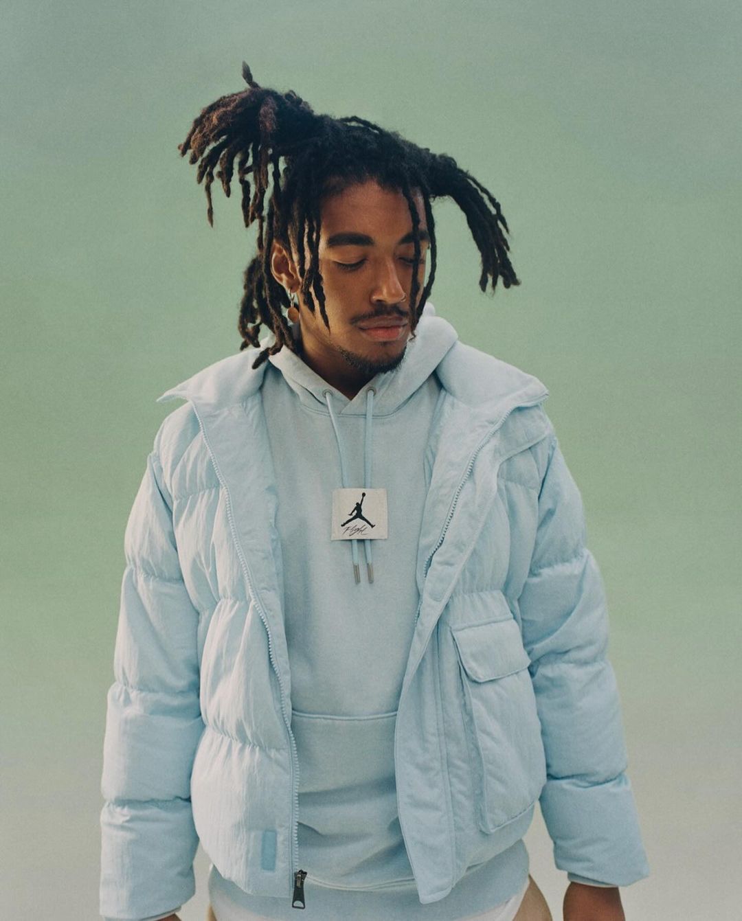 Person with dreadlocks wears a light blue puffer jacket and hooded sweatshirt indoors against a light green background in a portrait.