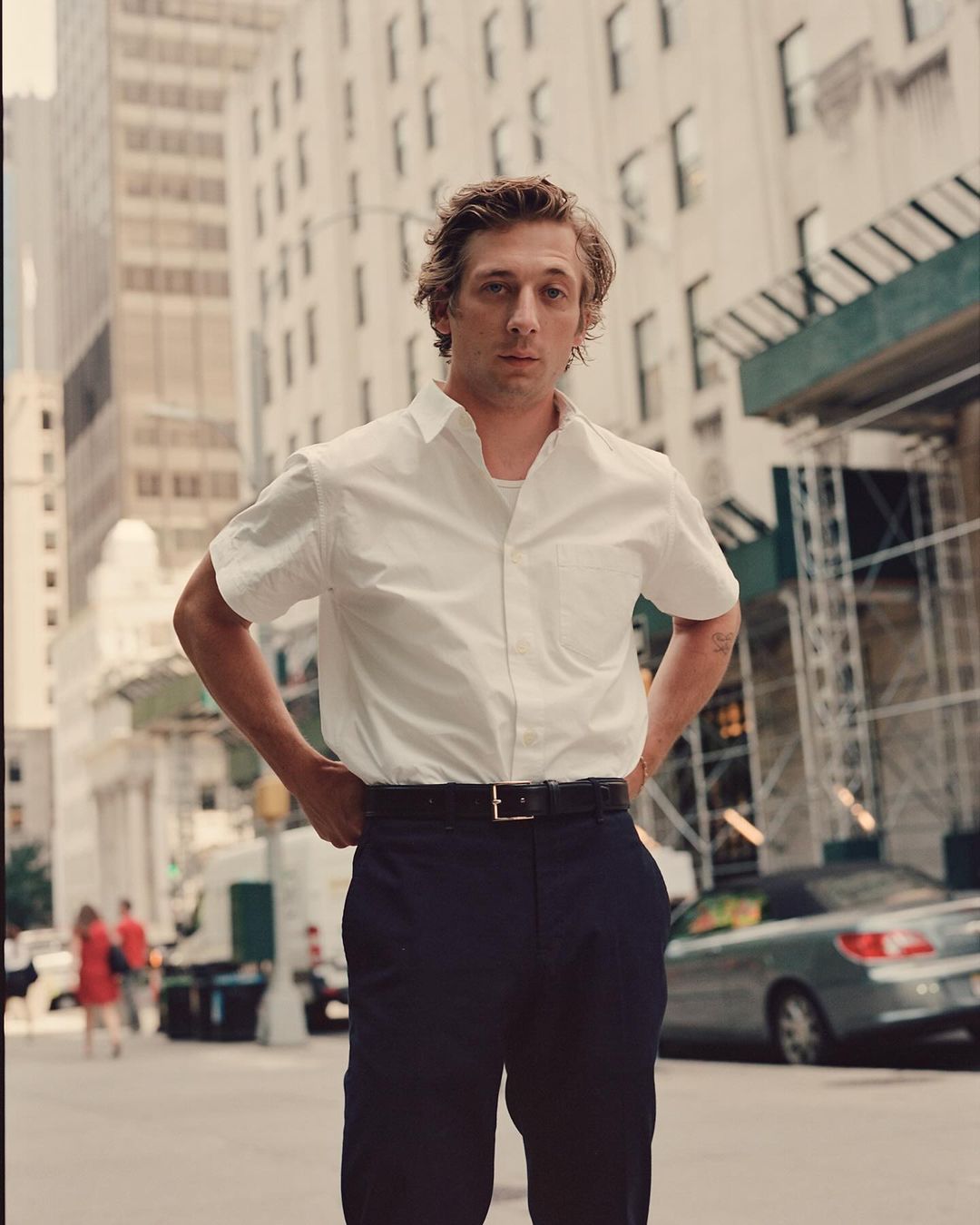 Man in white shirt and dark pants stands with hands on hips in front of tall buildings in an urban environment during daylight hours.