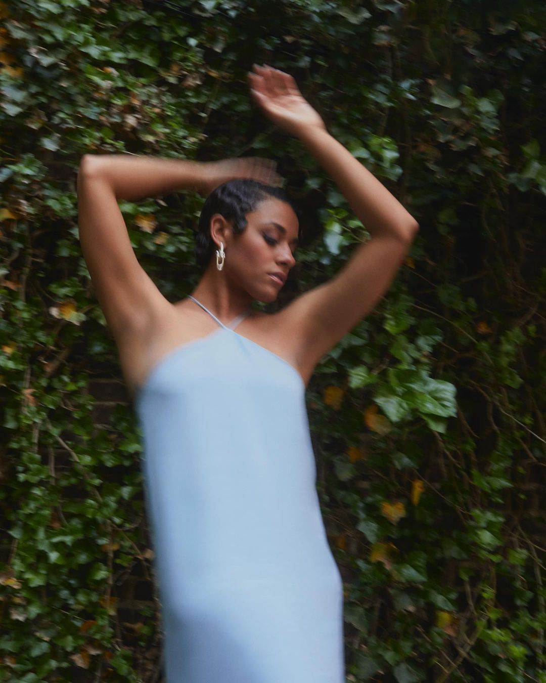 Woman in light blue dress poses with arms raised in front of ivy-covered brick wall, capturing a fashion-forward, nature-integrated aesthetic.