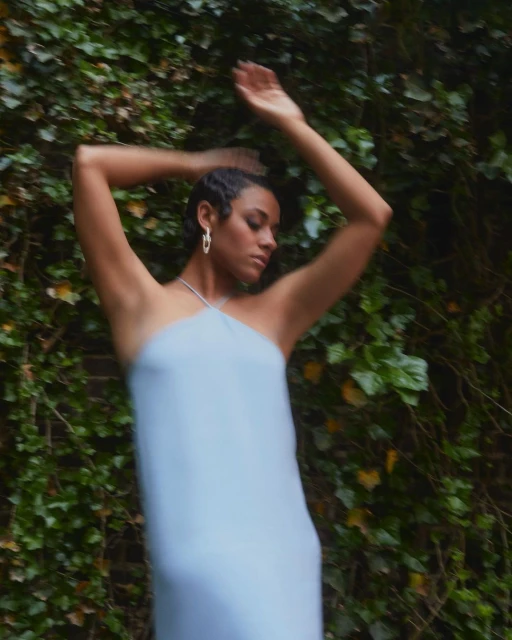 Woman in light blue dress poses with arms raised in front of ivy-covered brick wall, capturing a fashion-forward, nature-integrated aesthetic.