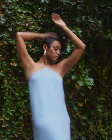 Woman in light blue dress poses with arms raised in front of ivy-covered brick wall, capturing a fashion-forward, nature-integrated aesthetic.