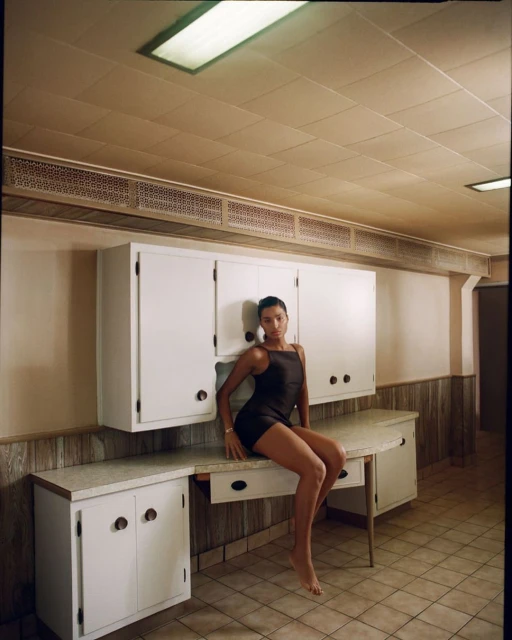 Woman in black dress sits on white countertop in a minimalist kitchen-like space with beige walls and tiled floor under fluorescent lighting.
