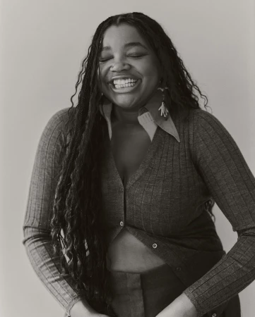 Black woman with braided hair laughing with closed eyes, wearing a buttoned ribbed cardigan against a plain background