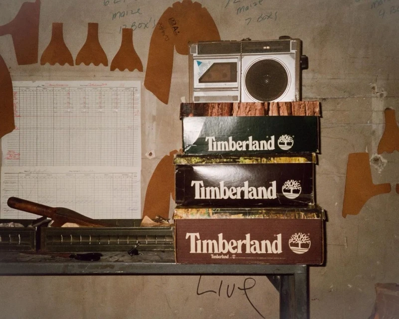 Stack of Timberland shoeboxes topped with a boombox on a metal table against a concrete wall adorned with hand-drawn notes and diagrams.
