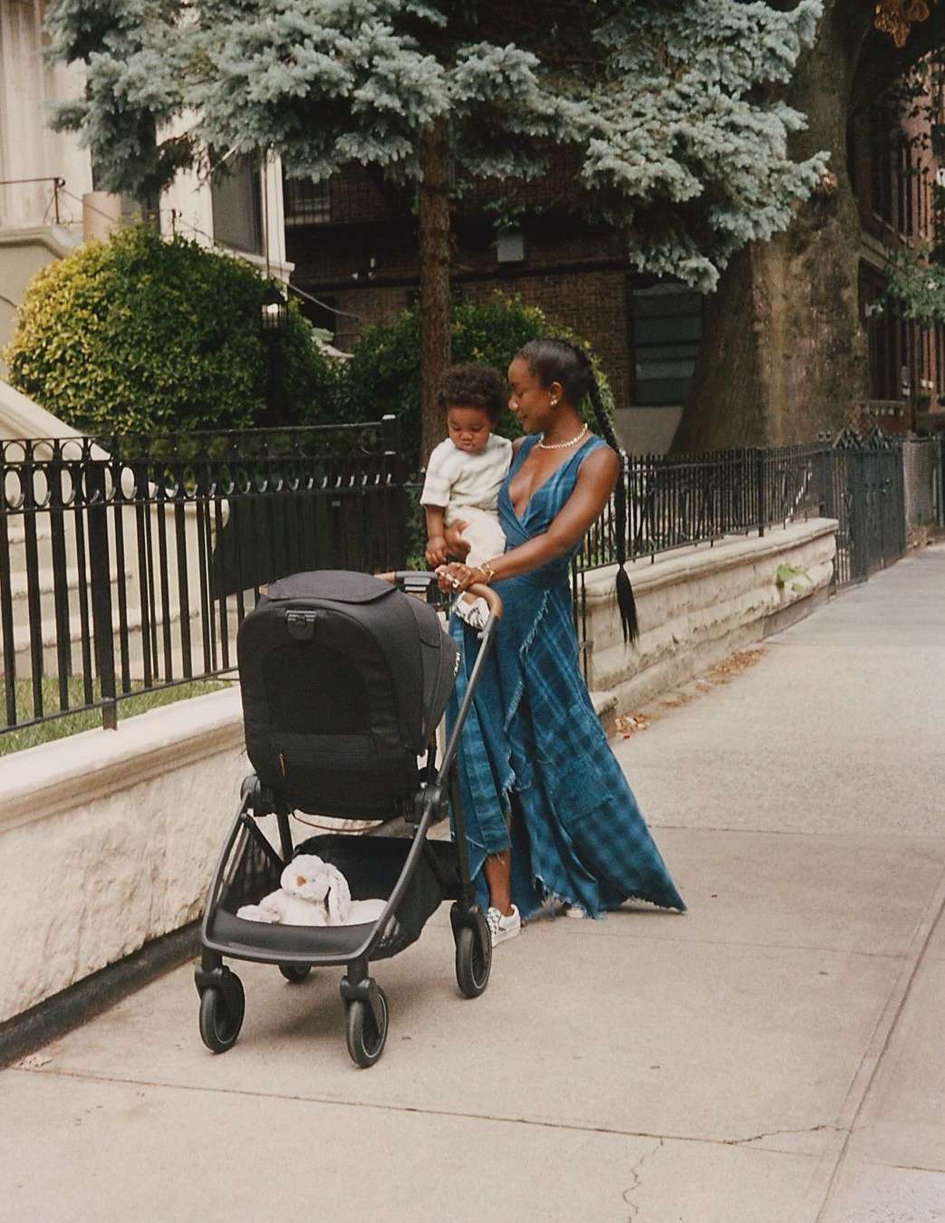 Woman walks on sidewalk with baby stroller and toddler in arms, set against buildings and sky, creating a family lifestyle scene.