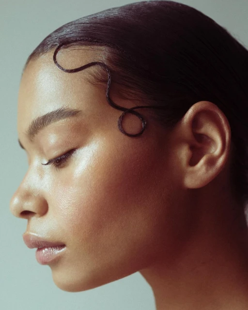 Profile portrait of a young adult woman with a dark complexion, slicked-back hair, and subtle makeup against a soft grey background.