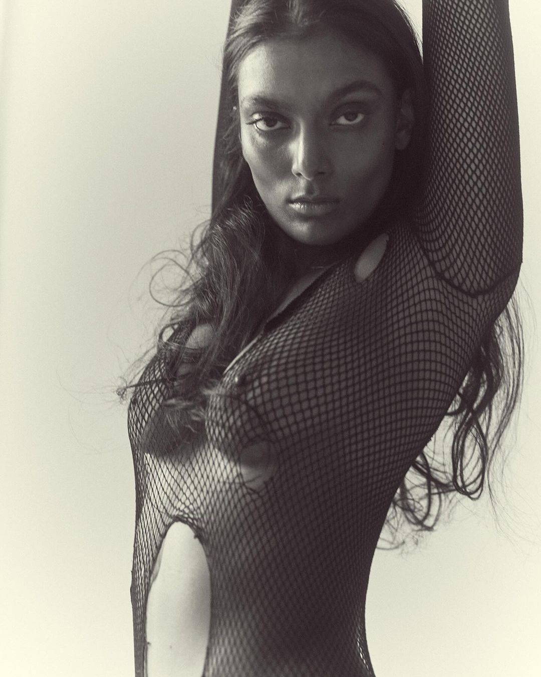 Striking black-and-white portrait of an adult female model in a torn fishnet top, posed against a plain white background.