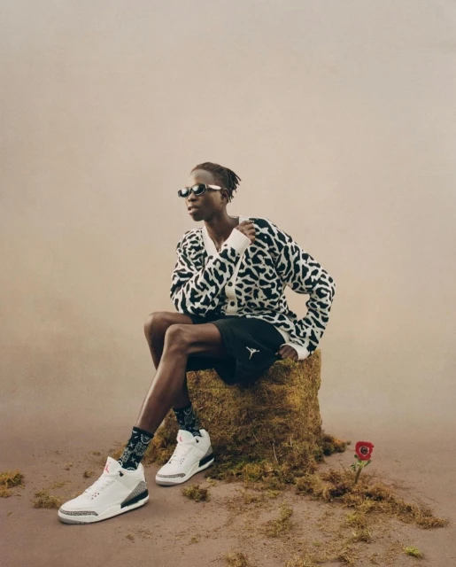 An adult wearing sunglasses and a patterned cardigan poses on a mossy tree stump next to a rose while showcasing white and gray sneakers.