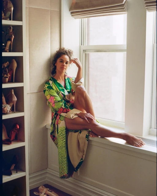 Woman in floral robe sits barefoot in window of interior room with shoe shelves. Indoor fashion portrait with natural light.
