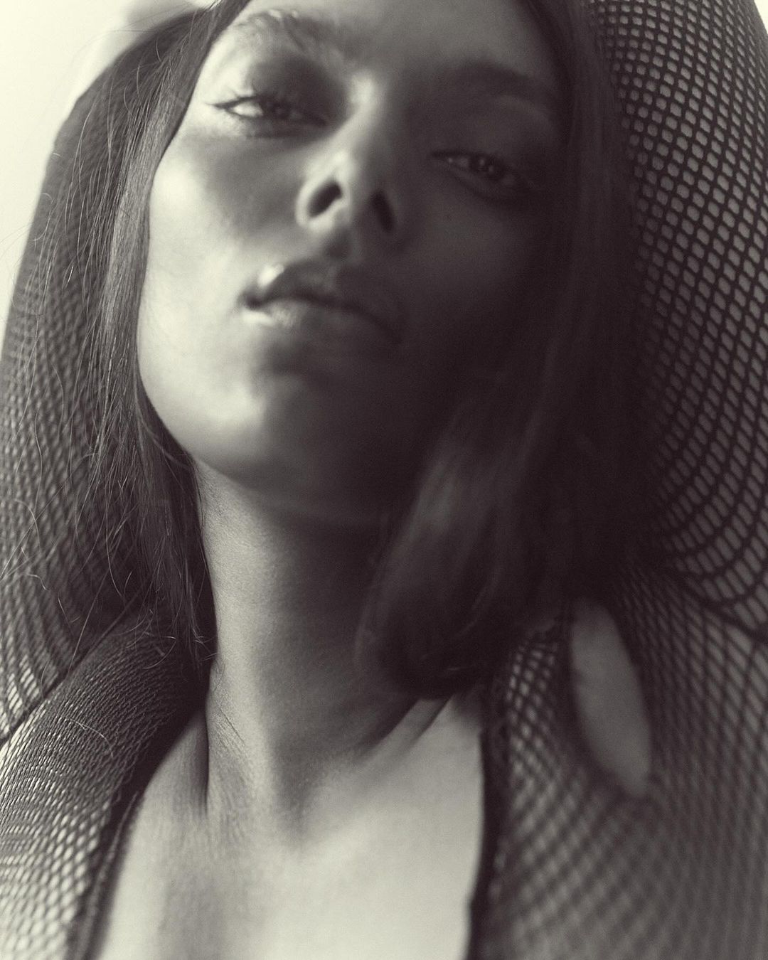 Monochrome close-up portrait of an adult female with dark hair and fishnet clothing, looking slightly upwards at the camera.