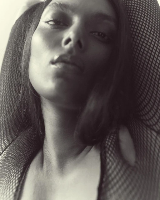 Monochrome close-up portrait of an adult female with dark hair and fishnet clothing, looking slightly upwards at the camera.