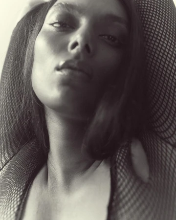 Monochrome close-up portrait of an adult female with dark hair and fishnet clothing, looking slightly upwards at the camera.