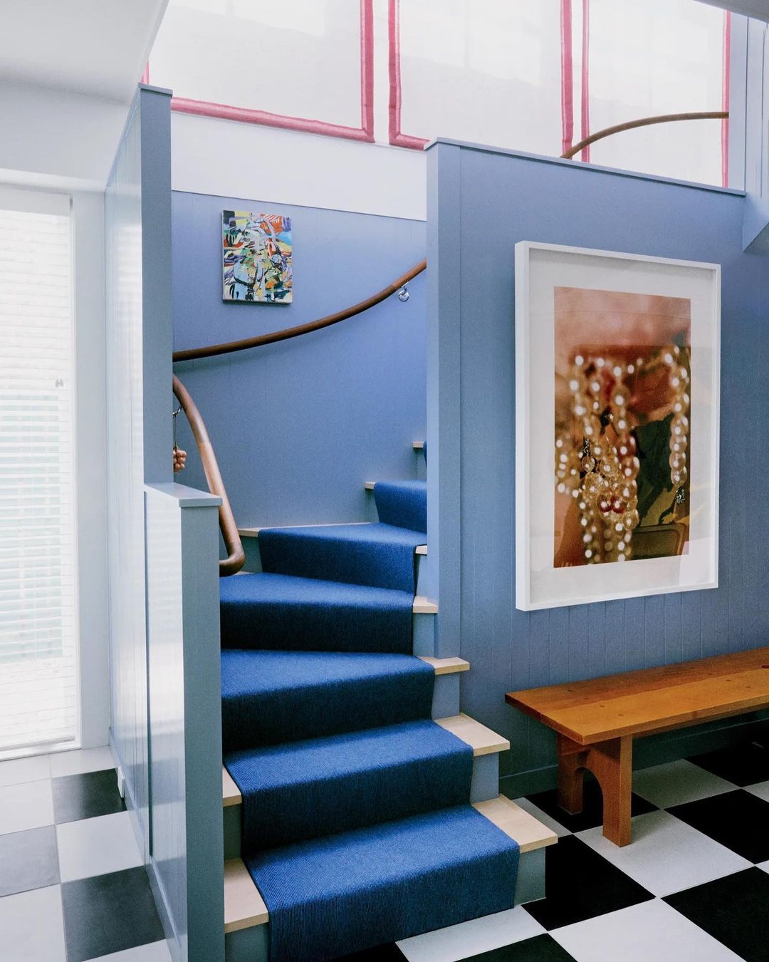 Interior view of a home featuring a blue carpeted staircase, light blue walls, a checkerboard tile floor, and artwork hanging on the walls.