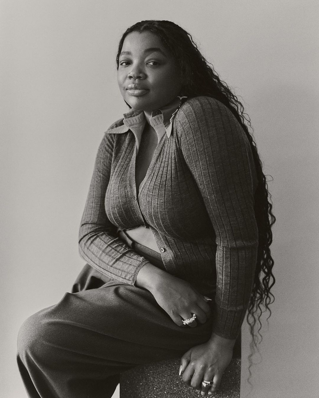 Portrait of Black woman with long wavy hair wearing a collared knit top and trousers sitting indoors in front of a neutral wall.