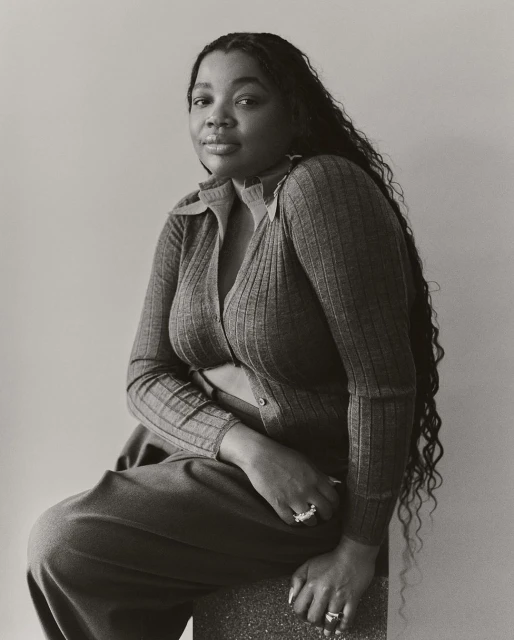 Portrait of Black woman with long wavy hair wearing a collared knit top and trousers sitting indoors in front of a neutral wall.